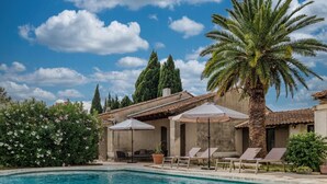 A swimming pool with lounge chairs and umbrellas, surrounded by greenery and a clear sky.
