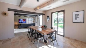 A dining area with a wooden table and metal chairs, a kitchen in the background, and a large window with a view of greenery.