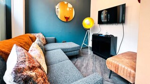 Una sala moderna con un sofá seccional gris, un espejo redondo, una lámpara de pie amarilla y un televisor de pantalla plana montado en la pared.
