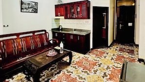 Un salon avec un tapis à motifs, un canapé en bois, une petite table et un coin cuisine doté de placards et d’un plan de travail.