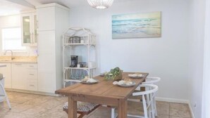 A dining area with a wooden table and white chairs, a white cabinet, and a hanging light fixture. - Panama City Beach -Beach House | Walk to Beach | Backyard Hangout | Sleeps 8 (Panama City Beach)