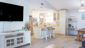 A modern kitchen with white cabinetry, a central island with stools, and a dining area with a wooden table. - Panama City Beach -Beach House | Walk to Beach | Backyard Hangout | Sleeps 8 (Panama City Beach)