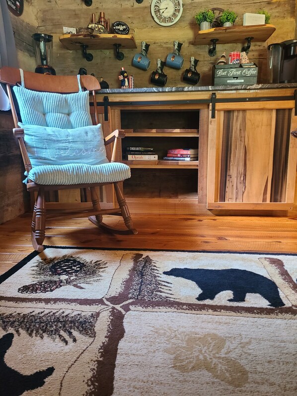 A wooden rocking chair with a striped cushion, a bookshelf with books, and a coffee station with mugs and a coffee grinder.