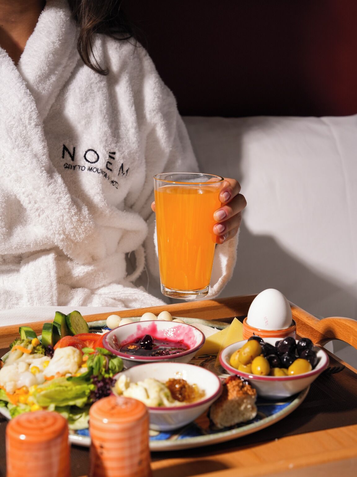 Une personne vêtue d’une robe blanche tient un verre de jus d’orange et porte un plateau contenant divers aliments, notamment une salade, des œufs et des olives.