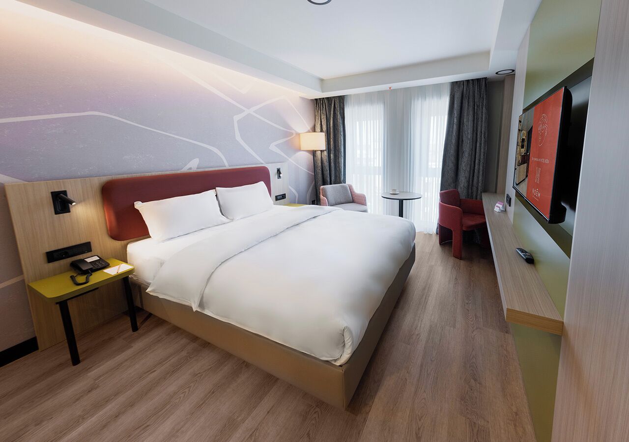 A hotel room with a large bed, a bedside table with a phone, a small table with a chair, and a TV mounted on the wall.
