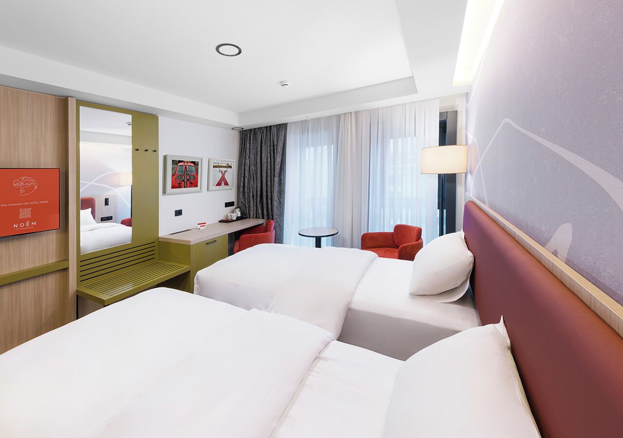 Une chambre d’hôtel avec deux lits, un bureau, une chaise, une petite table et une grande fenêtre avec des rideaux.