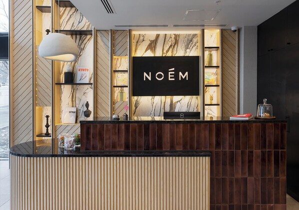 Un mostrador de recepción con un fondo de pared de mármol, un letrero que dice "NOËM" y un diseño moderno con paneles de madera verticales.