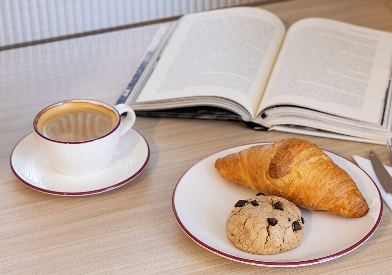Un croissant, una galleta y una taza de café sobre una mesa con un libro abierto.