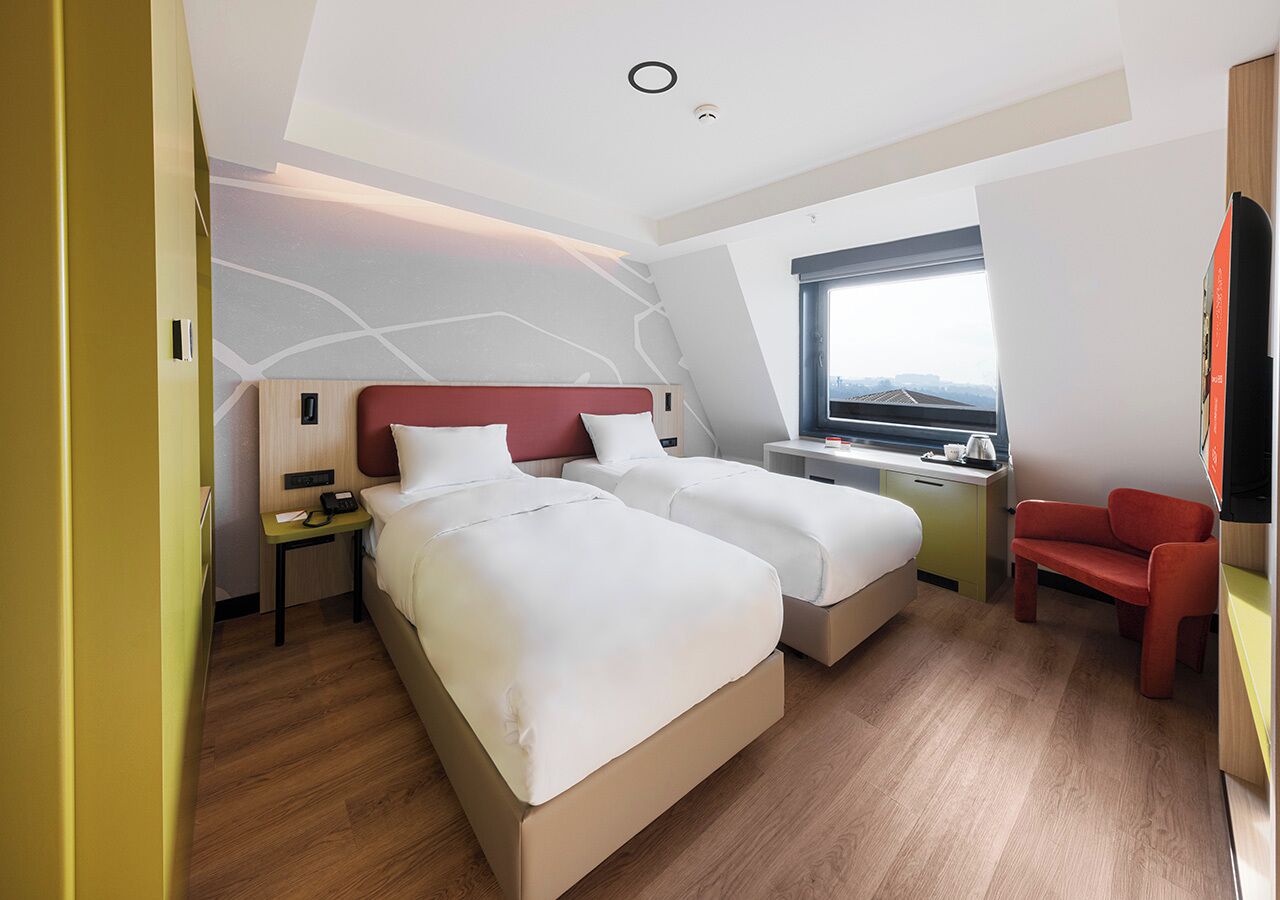 A modern hotel room with two beds, a desk, a chair, and a large window.