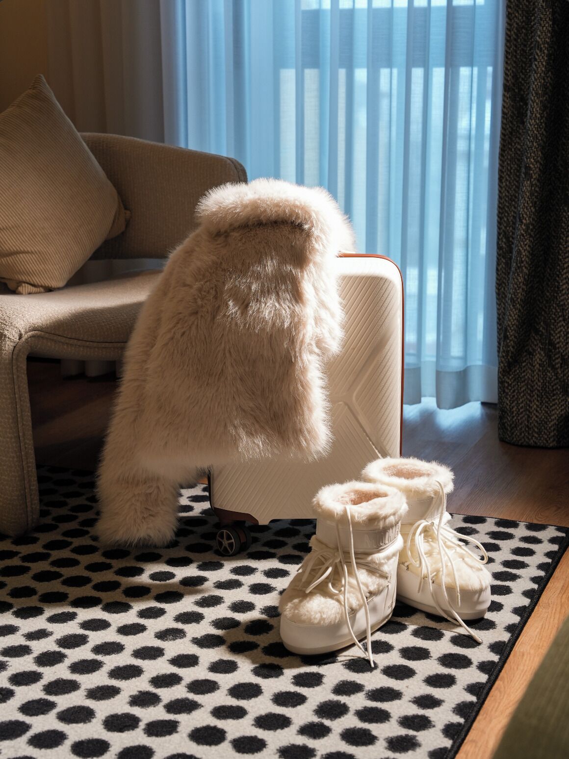 A fluffy white dog sitting on a chair, a pair of white boots on a patterned rug, and a window with sheer curtains.