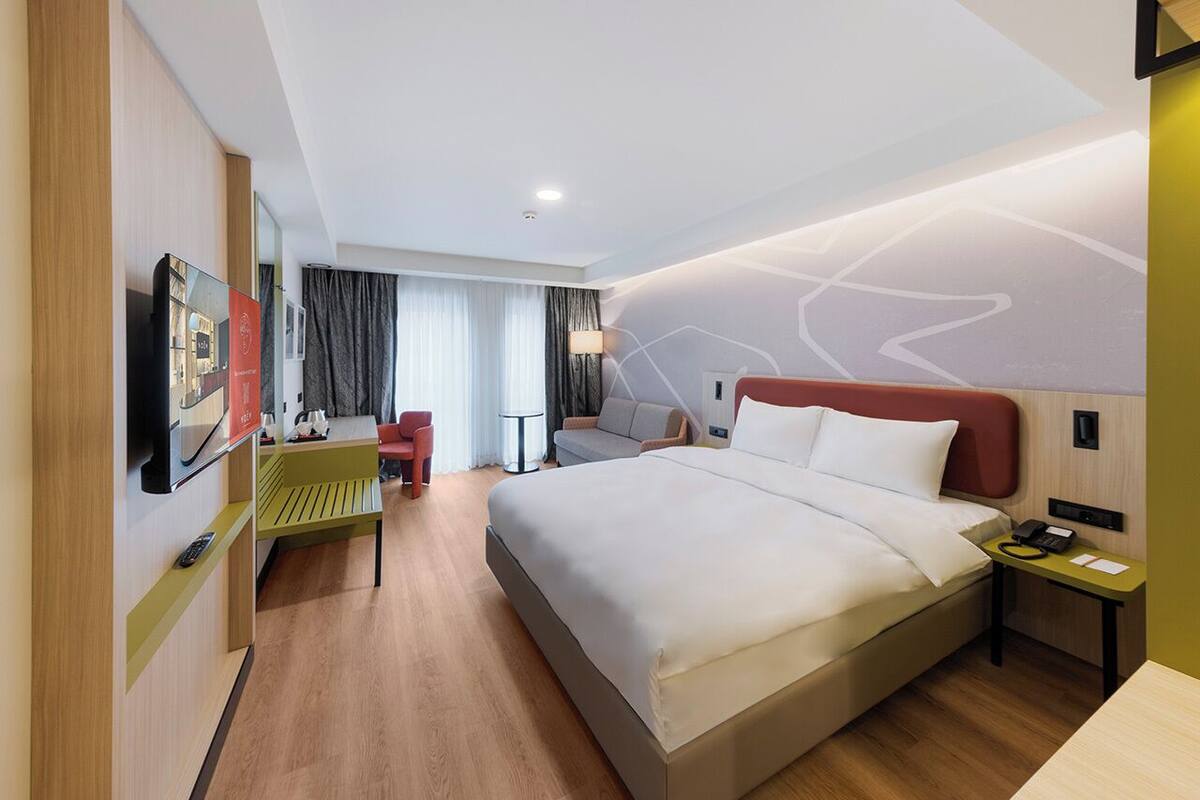 A modern hotel room with a large bed, a desk, a chair, and a TV mounted on the wall.
