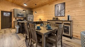 Un interior de madera con una mesa de comedor preparada para cuatro, una zona de cocina con electrodomésticos modernos y un gran cuadro abstracto en la pared.