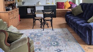 A cozy living room with a wooden floor, a patterned rug, a green armchair, a blue sofa, and a kitchen area with a microwave and kettle. - Apartment-sleeps2-parking-garden-patio (Castleisland)