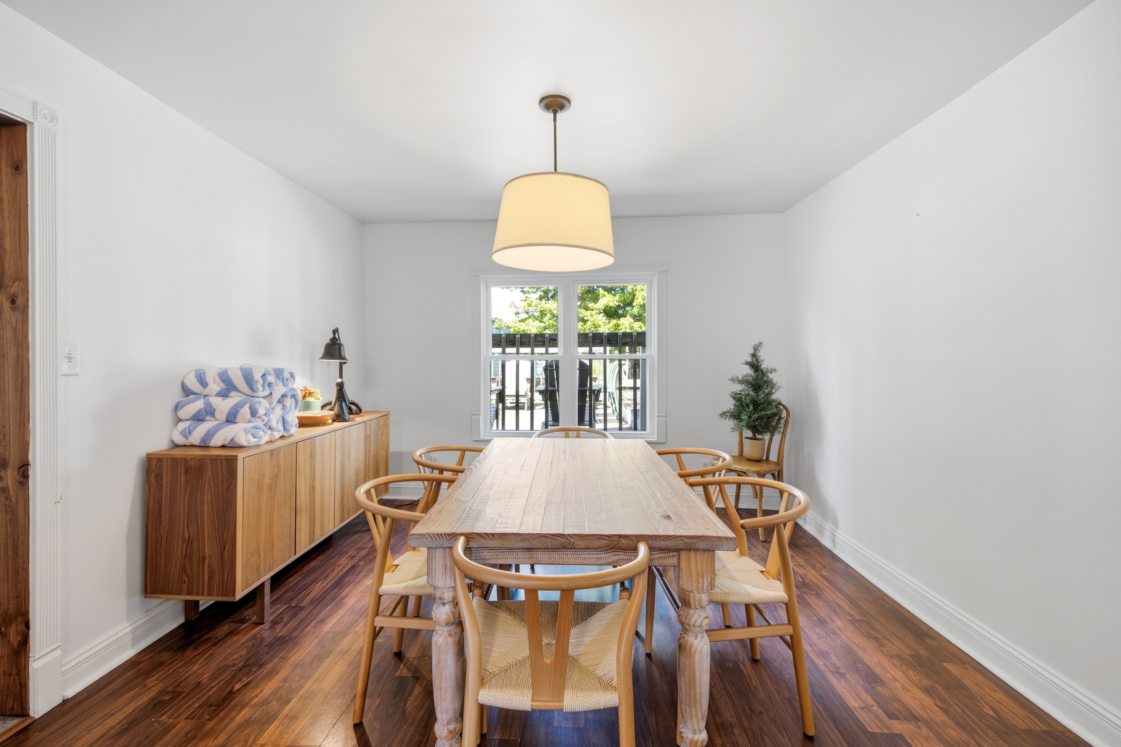 A dining room with a wooden table and chairs, a hanging light fixture, and a view through a window.