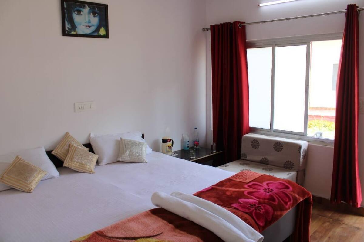A hotel room with a bed, pillows, a bedside table with a lamp, a window with red curtains, and a view of the outdoors.