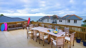 Un espace extérieur abrité, aménagé pour les repas, avec une table et des chaises en bois, entouré d’une clôture en bois, et offrant une vue sur un plan d’eau et des maisons.