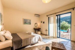 Quarto com cama, mesa de cabeceira com abajur, pintura na parede e vista para uma piscina e montanhas pela porta aberta.