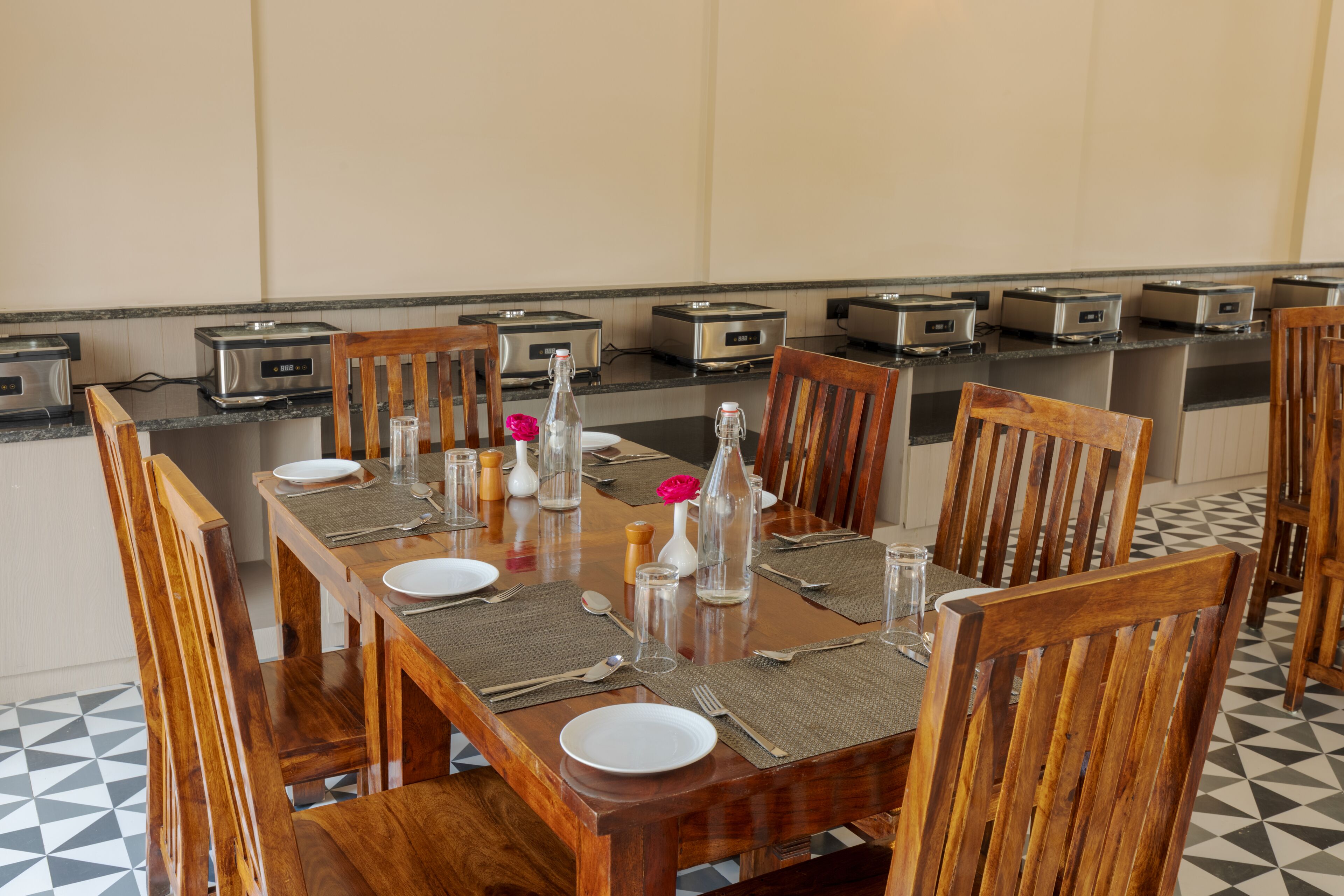Un coin repas avec une longue table en bois, dressée pour six personnes, agrémentée de grille-pains, de salières et de poivrières, et d’un vase contenant une fleur rose.