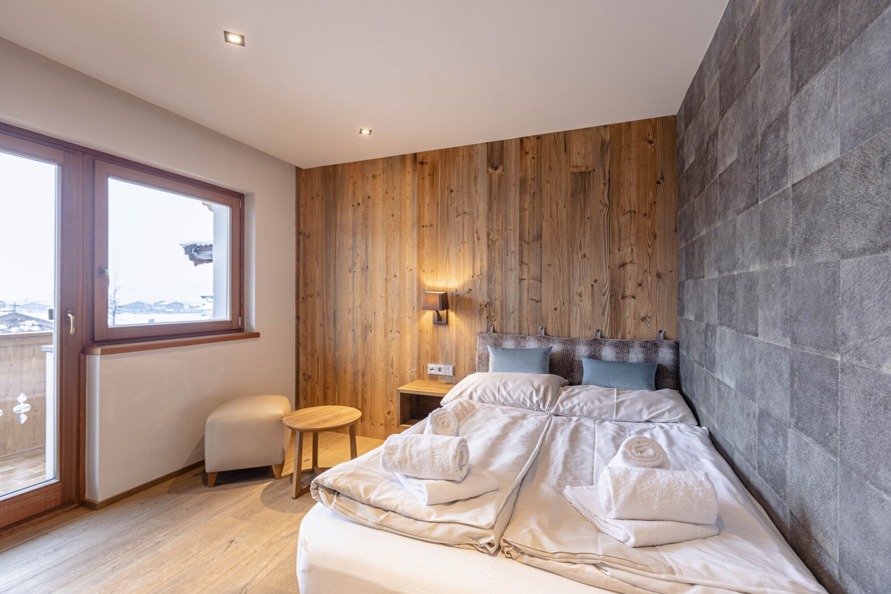 A bedroom with a wooden wall, a bed with white linens, a small wooden stool, and a nightstand with a lamp.