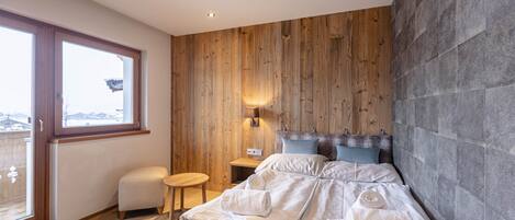 A bedroom with a wooden wall, a bed with white linens, a small wooden stool, and a nightstand with a lamp.