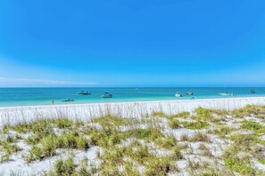 Una playa de arena blanca y vegetación escasa, con barcos en el agua y un cielo azul despejado.