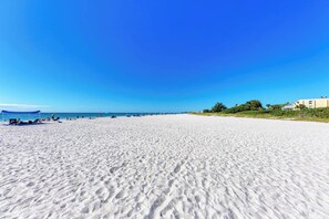 Una amplia playa de arena, cielo azul despejado, algunas sombrillas y edificios a lo lejos.