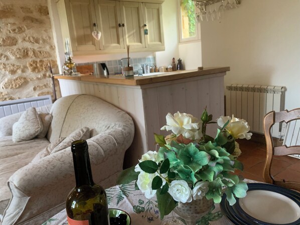 Ein gemütliches Wohnzimmer mit einer Couch, einem Tisch mit einer Flasche und Blumen, und einer Küchenzeile im Hintergrund.