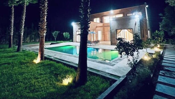 Una casa moderna con piscina, rodeada de palmeras e iluminada durante la noche.