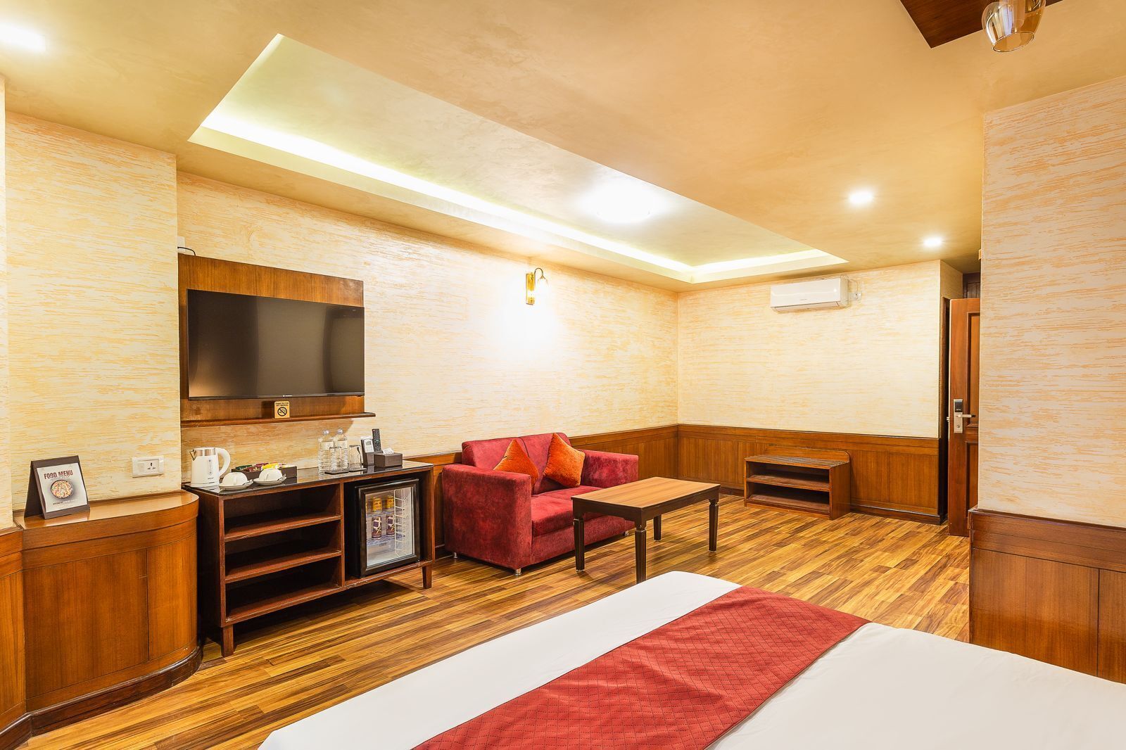 A hotel room with a wooden floor, a red sofa, a small table, a TV mounted on the wall, and a minibar.