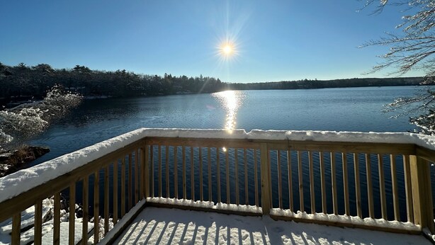 Una terraza de madera con barandillas cubiertas de nieve, con vistas a un lago tranquilo bajo un cielo despejado y con el sol reflejado en el agua.