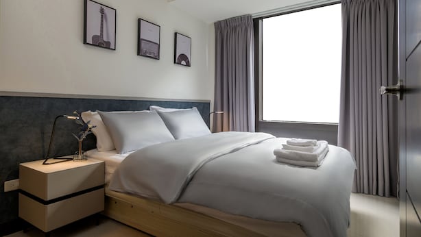 A neatly made bed with white linens, a bedside table with a lamp, and a window with curtains. - Taipei Main Station Modern Comfort 2B1b Living, Dining, Kitchen (Taipei City)