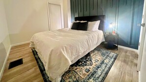 A bedroom with a bed, a nightstand, a wall-mounted shelf, a door, and a window.