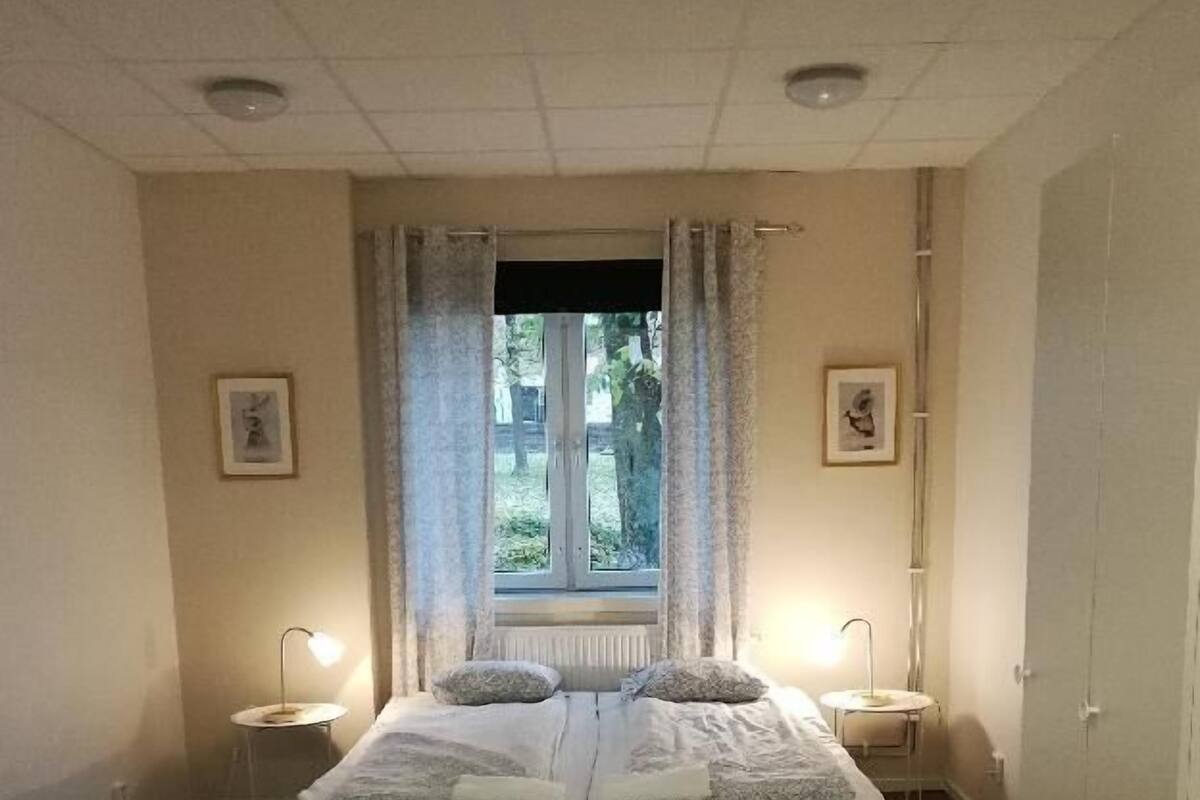 Quarto com cama de casal, duas mesas de cabeceira, janela com cortinas, radiador e tapete estampado.