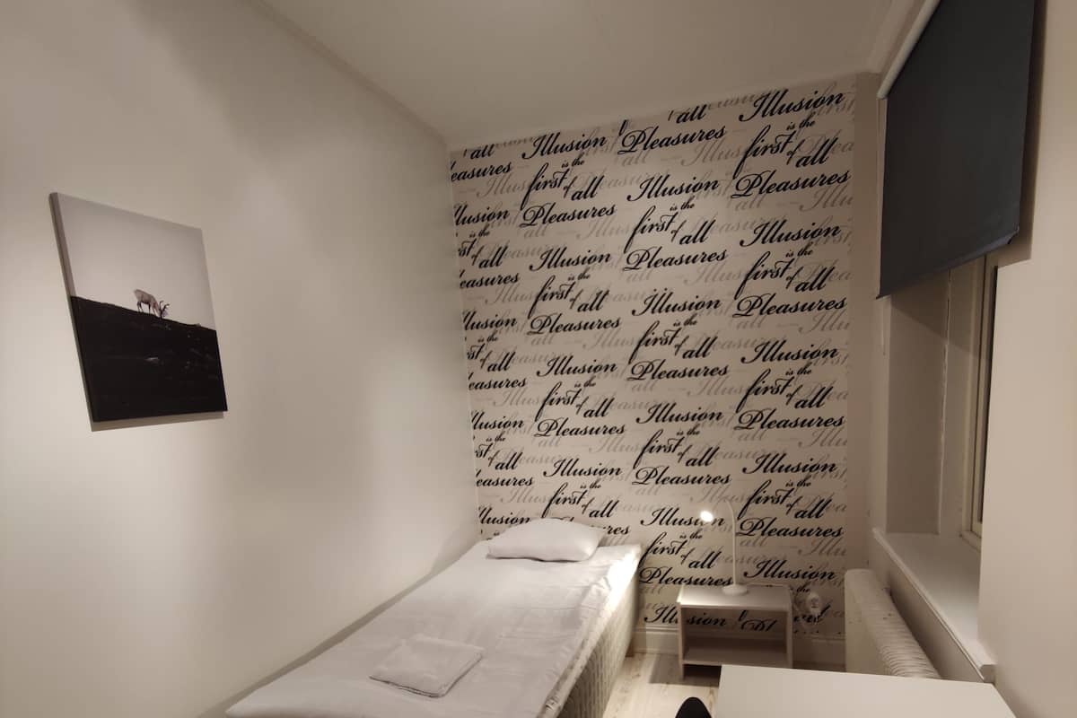 Quarto pequeno e bem organizado, com uma cama de solteiro, uma escrivaninha e uma parede com texto repetido.