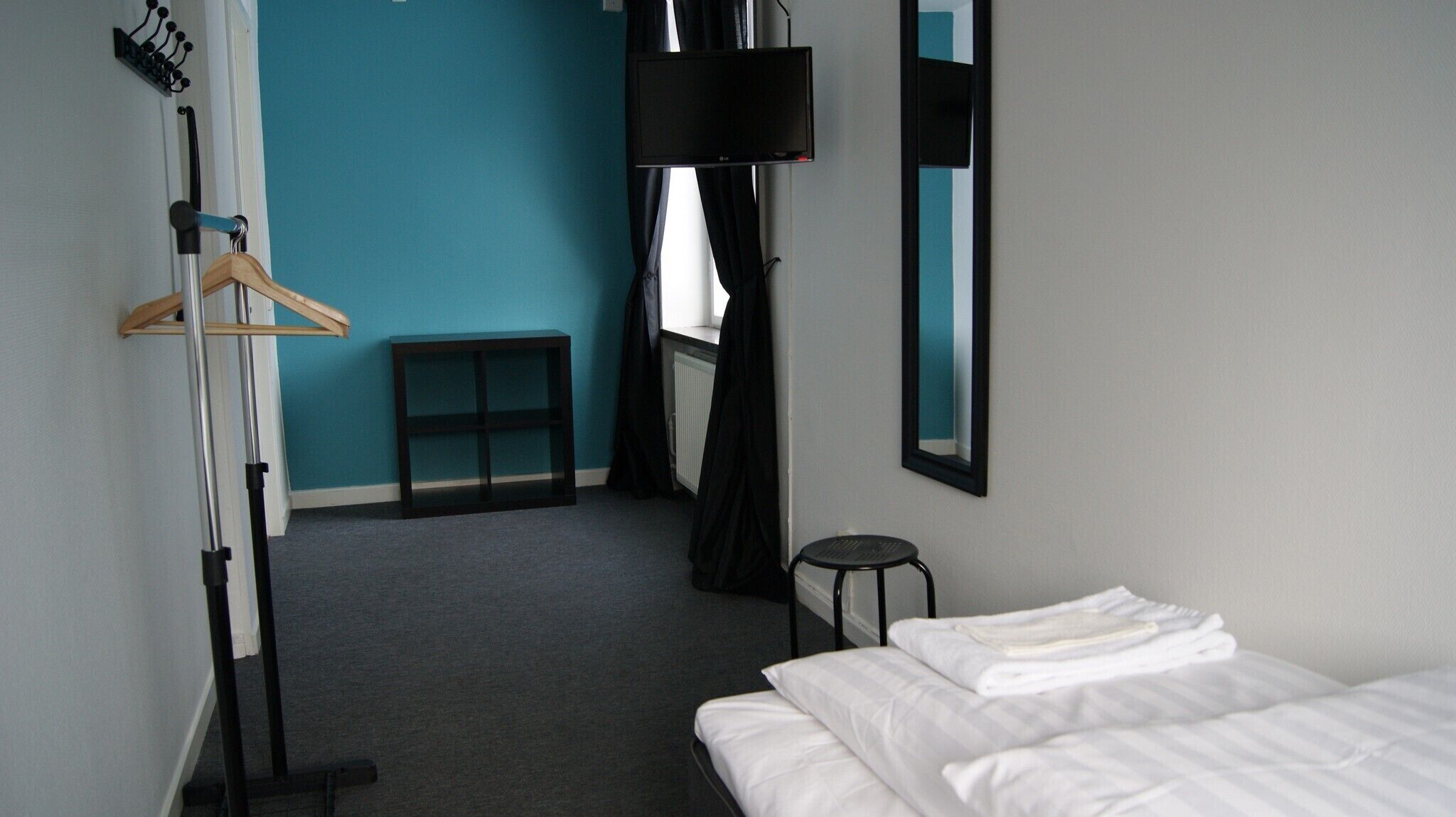 A hotel room with a bed, a small table, a hanging TV, and a coat rack.