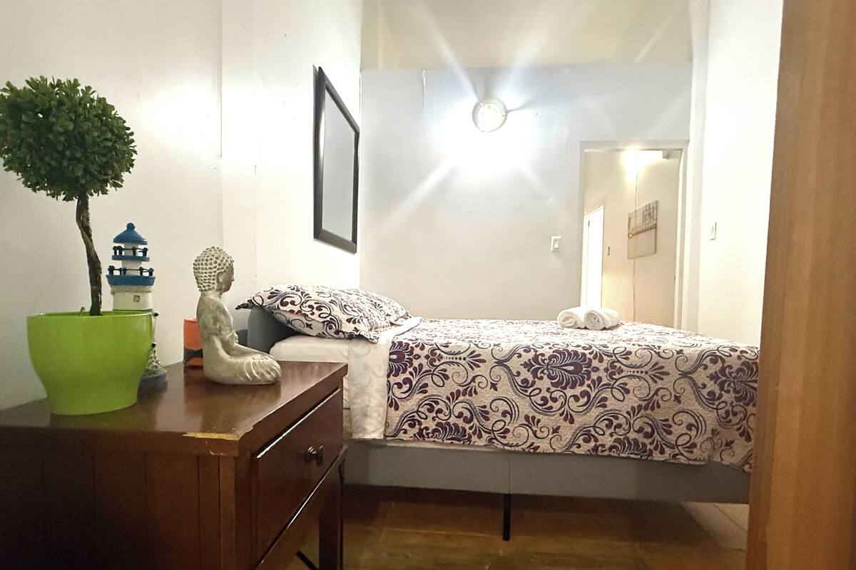 A bedroom with a bed, a desk, a potted plant, a Buddha statue, and a ceiling fan.