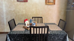 Een eetruimte met een tafel voor vier personen, stoelen, een koelkast en twee schilderijen aan de muur.