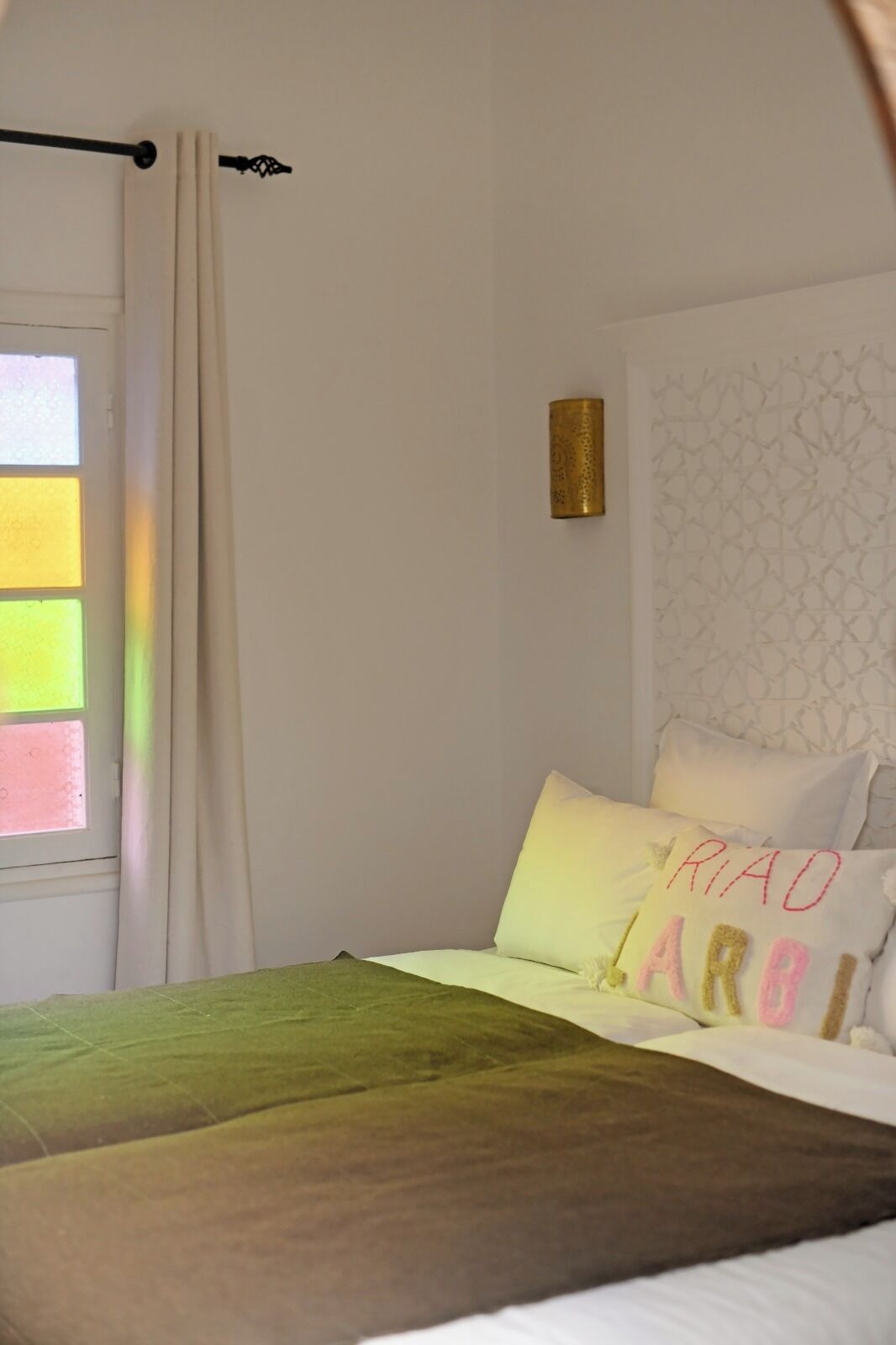 A bedroom with a bed, a window with curtains, a wall-mounted lamp, and a decorative pillow.