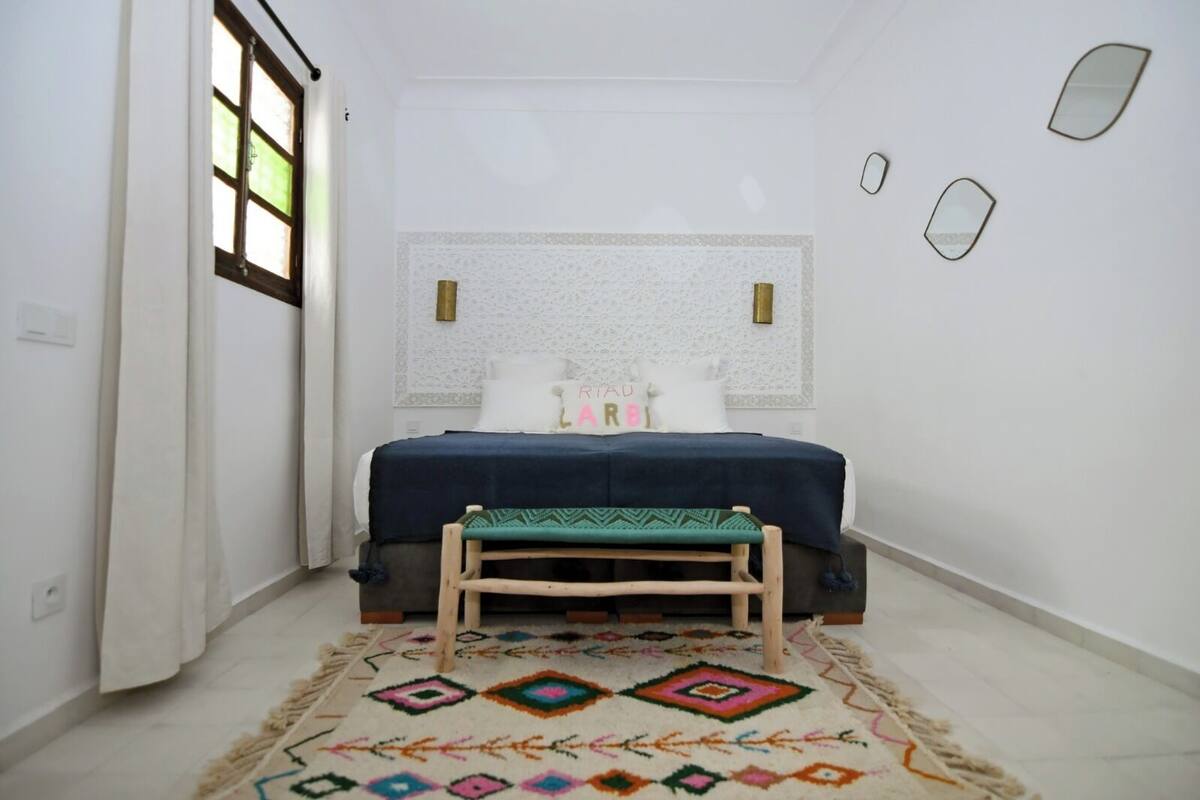 A bedroom with a bed, a bench, a patterned rug, and a window with curtains.