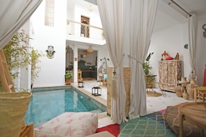 A spacious indoor pool area with a view of a kitchen and living space, featuring a hanging chair, potted plants, and a decorative cabinet. - Riad larbi khalis  (Marrakech)