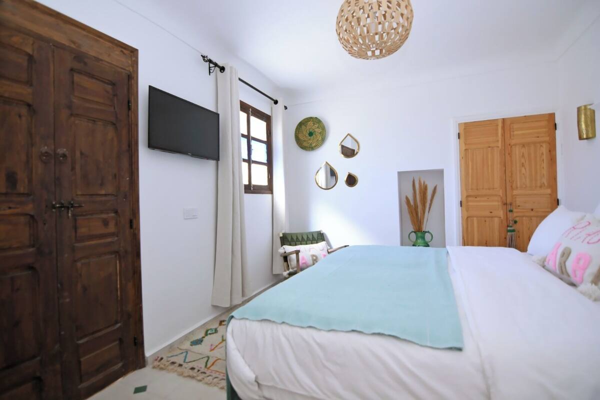 A bedroom with a bed, wooden wardrobe, a TV mounted on the wall, and a hanging light fixture.