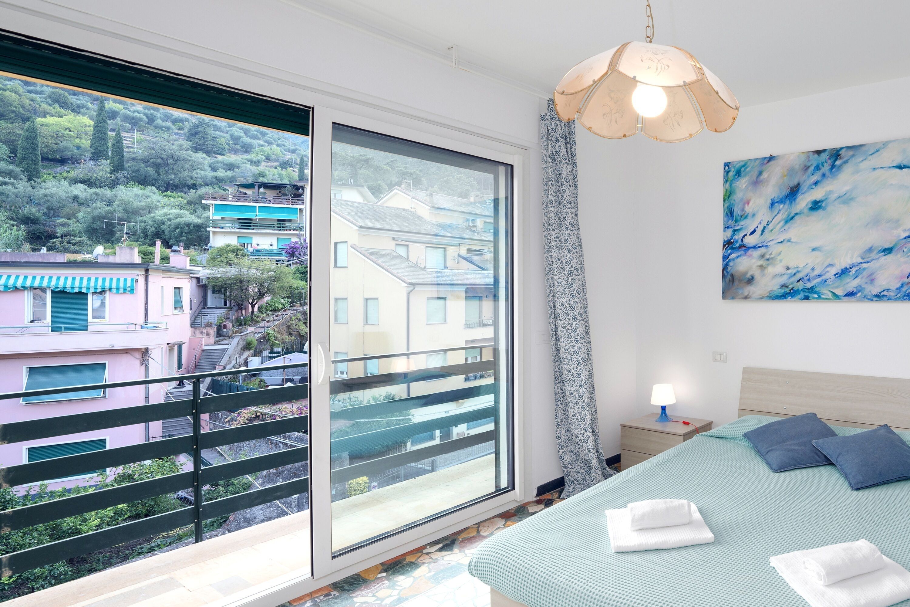 Une chambre avec une grande fenêtre, un lit avec une literie bleue, une table de chevet avec une lampe et un tableau accroché au mur.