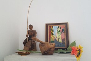 A wooden statue of a seated figure holding a staff, a rustic wooden bowl, and a framed painting of colorful buildings on a white surface.