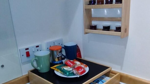 Una kitchenette compacta con estufa, un estante de madera con frascos, una taza y un plato con comida.
