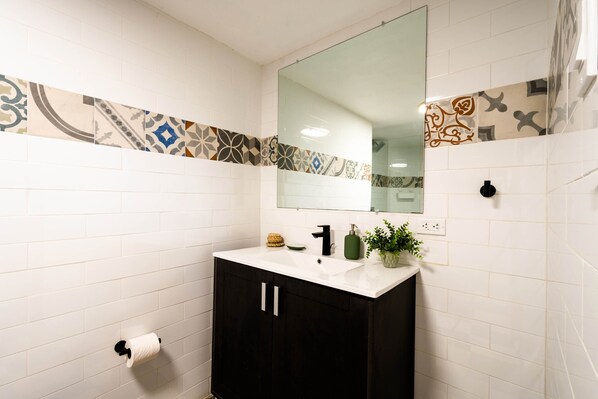 A bathroom with white tiled walls, a black cabinet, a mirror, and a patterned tile border.