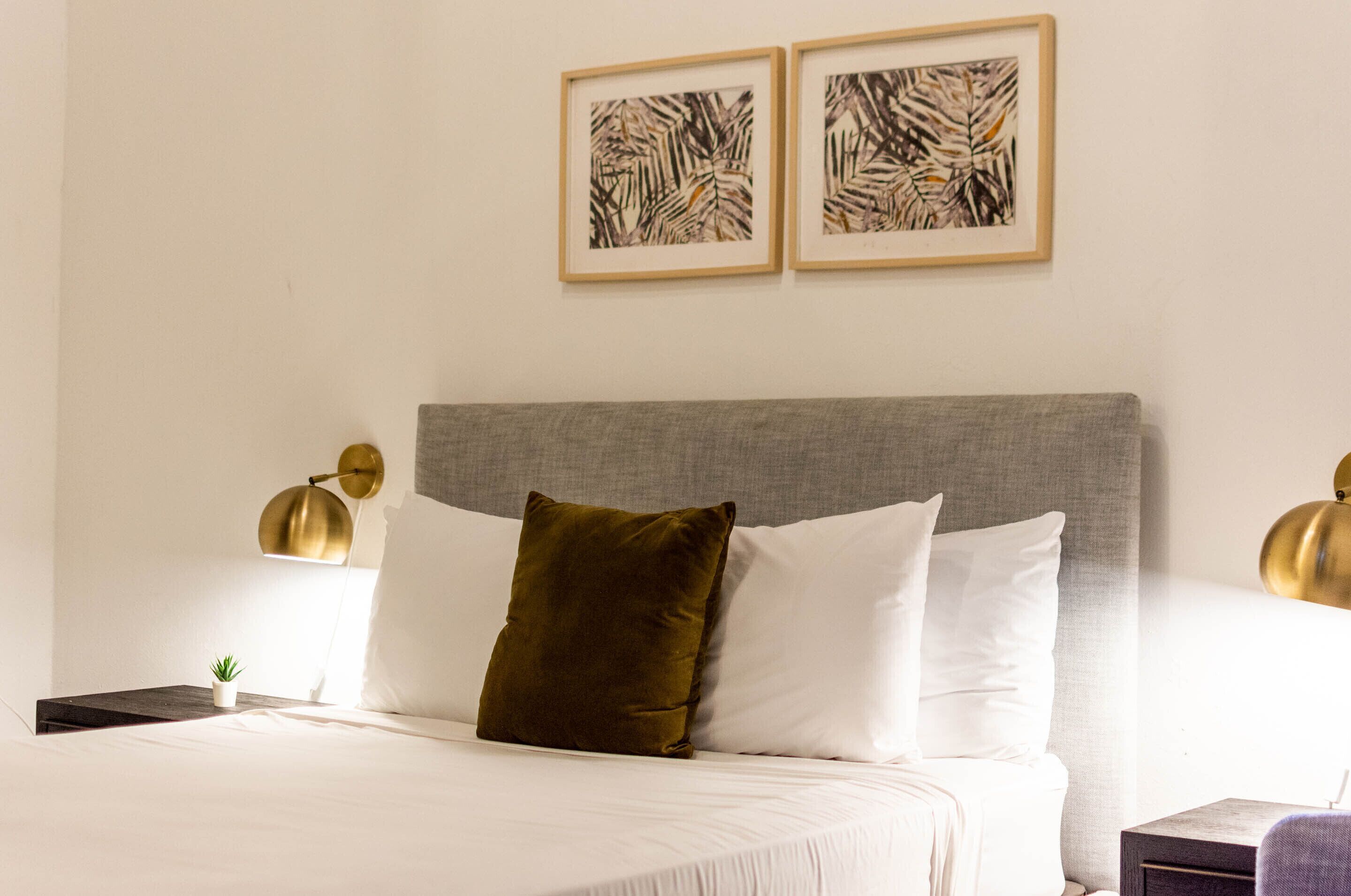 Ein ordentlich bezogenes Bett mit weißen und grünen Kissen, einem grauen Kopfteil und zwei gerahmten Kunstwerken an der Wand.