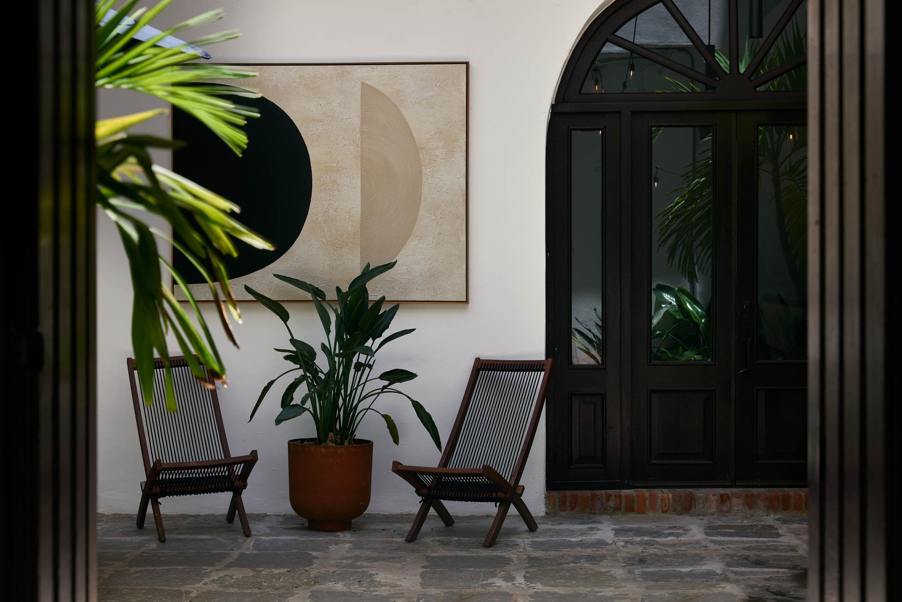 A room with a large abstract painting, two wooden chairs, a potted plant, and a view through a window.