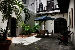 Une cour avec une table et des chaises, entourée de plantes en pot et agrémentée d’un parasol bleu.