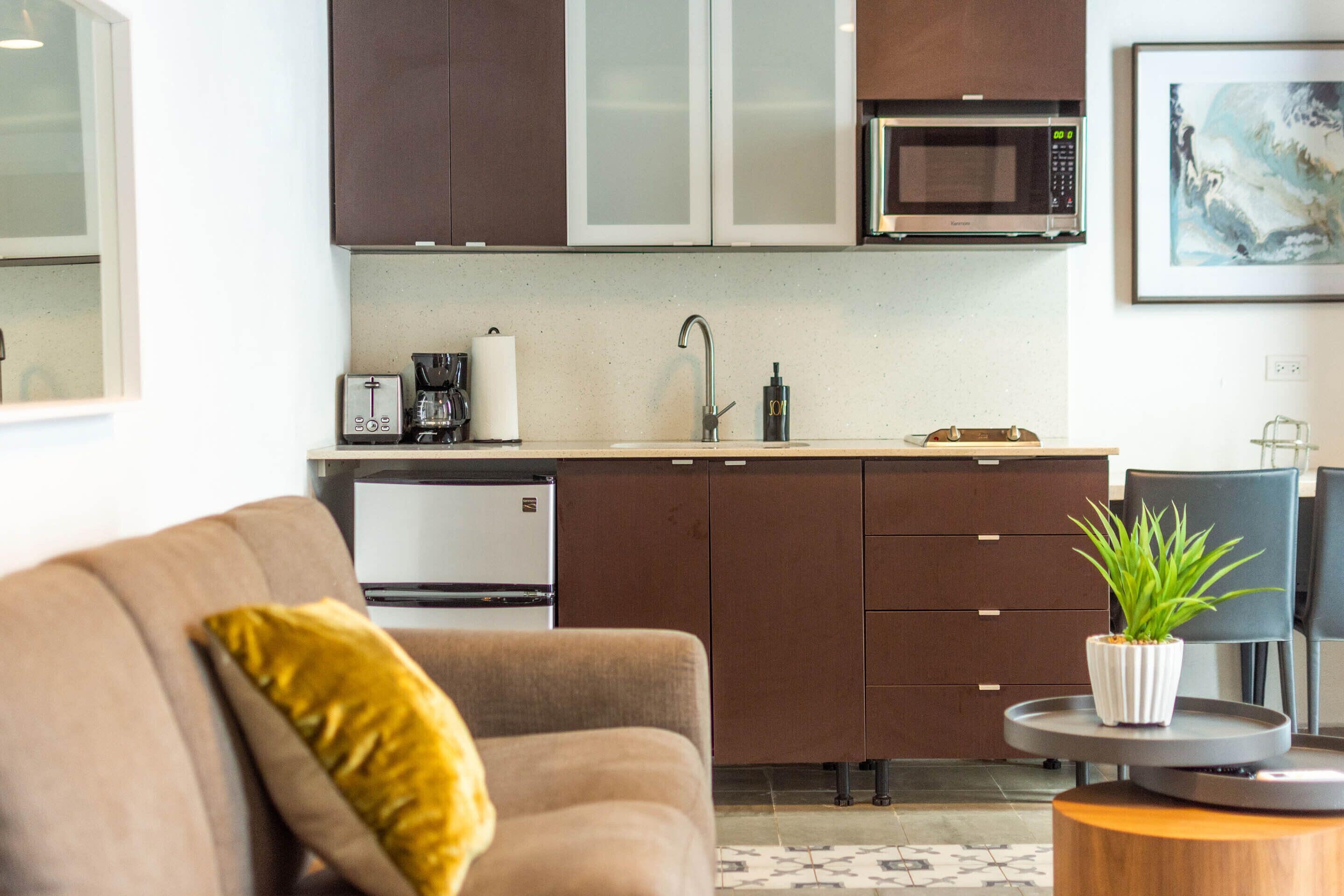 Cocina moderna con microondas, cafetera y lavabo. Hay un comedor con mesa y sillas, y un sofá con cojín.