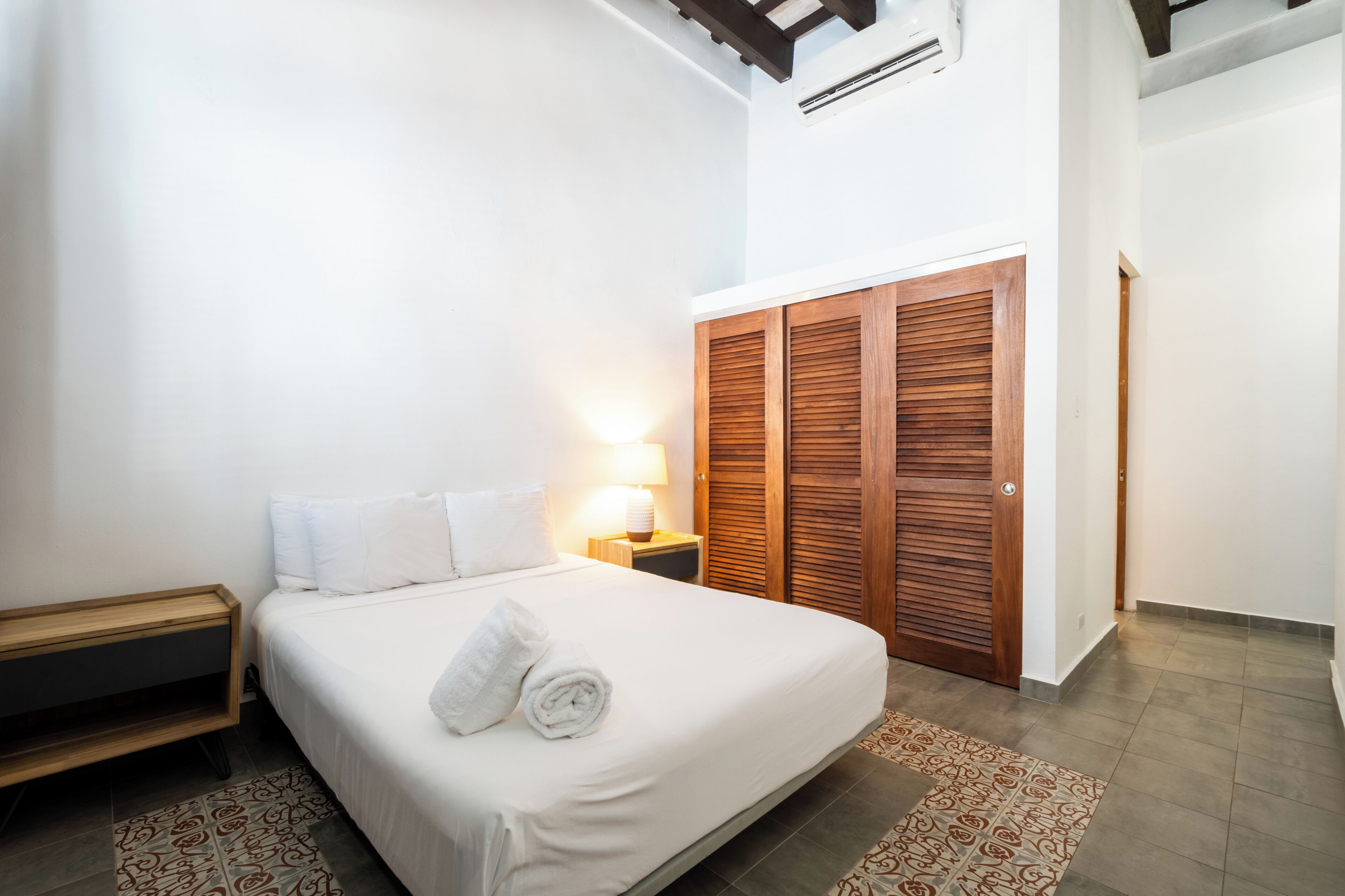 A hotel room with a bed, a nightstand, a lamp, and a wooden wardrobe.